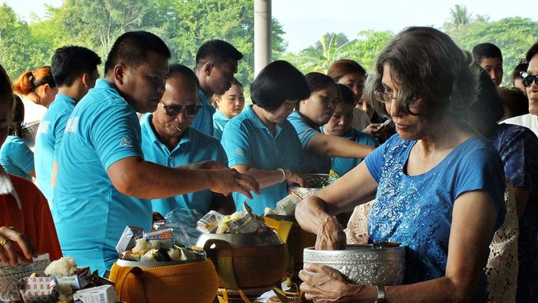 A Indian American woman mixes food in a pot standing up with many of her Thai friends. They are all wearing blue