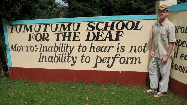 A young, white man stands outside next to the gate of TumuTumu School for the Deaf in Kenya..