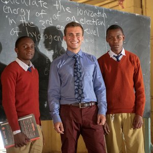 A white male Peace Corps Volunteer stands between two high school students in Tanzania. They are all dressed in uniforms.