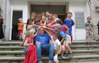 A male Volunteer sits outside on steps while a large group of children laugh and place their hands on his head.