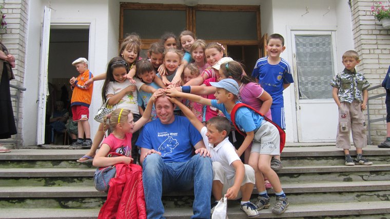 A male Volunteer sits outside on steps while a large group of children laugh and place their hands on his head.
