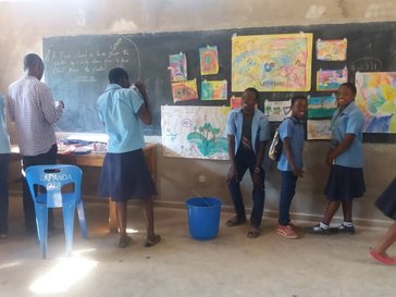 Students in blue uniforms stand at the front of the room looking at their paintings displayed on the black board