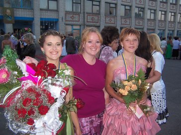 Three woman pose together with flowers in front of a school for their graduation celebration.