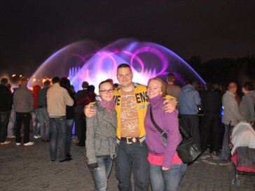Two women stand with a man posing in front of a crowd of people viewing a fountain at night in Vinnytsia.
