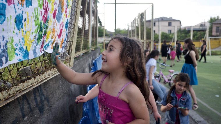 A young girl, hand covered in blue paint, adds her hand print to piece of paper with other colorful hand prints.