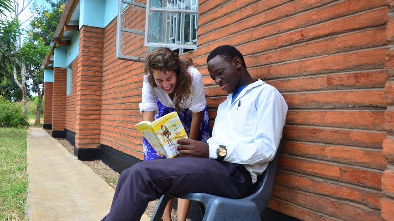 A female Volunteer works with a male student outside, with the help of a yellow book.