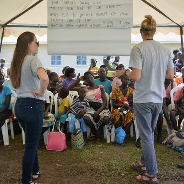 A male and female Volunteer stand with a male Ugandan counterpart and deliver a lesson about malaria to a group of about twen
