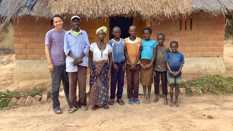 A Peace Corps Volunteer stands with his Zambian host family outside their home.