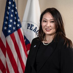 An Asian American woman stands smiling in front of an American flag for a professional head shot.