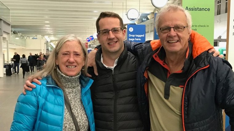 A young white male stands between his mother and father outside. All three smile.
