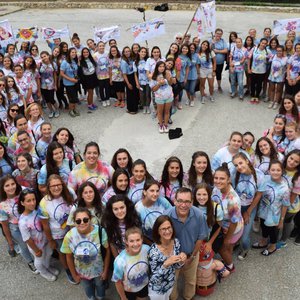 An overhead shot of a group of teen girls stand in the shape of a heart. They are wearing tie-dye shirts.