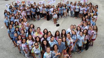An overhead shot of a group of teen girls stand in the shape of a heart. They are wearing tie-dye shirts.
