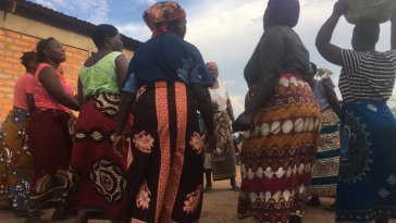 Malawian women stand in a cirlce wearing colorful chitenje cloth skirts and carrying buckets on their heads.