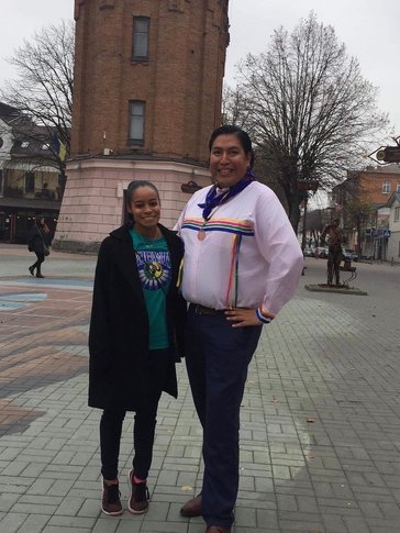 Two people of Indigenous heritage stand outside in their traditional clothing.