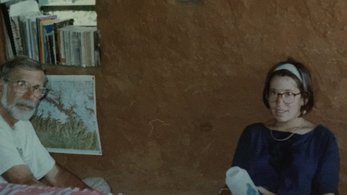 An older white man and young white woman sit together on the ground in front of a mud wall