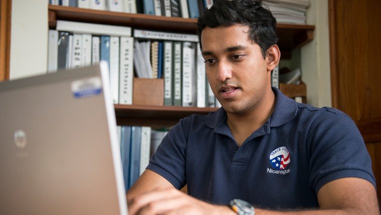 A young man of Color in a Peace Corps branded shirt types on a laptop.