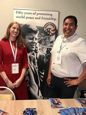 A man of Indigenous heritage and a white woman stand next to each other in front of a Peace Corps banner.