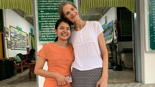 A tall, middle aged white woman and a young, short woman from Myanmar stand side by side, smiling and embracing..