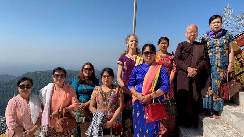 A large group of people in colorful, traditional Myanmar dress stand outside on a cliff.