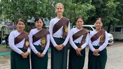 Five middle aged women stand in a row, outside, in traditional Burmese green and white outfits.