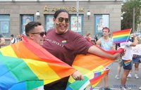 A man with a Native background wraps a rainbow flag around himself and a friend, smiling.