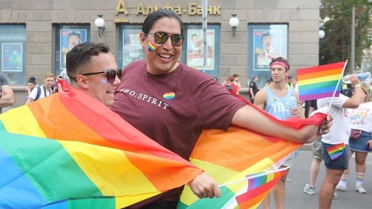 A man with a Native background wraps a rainbow flag around himself and a friend, smiling.