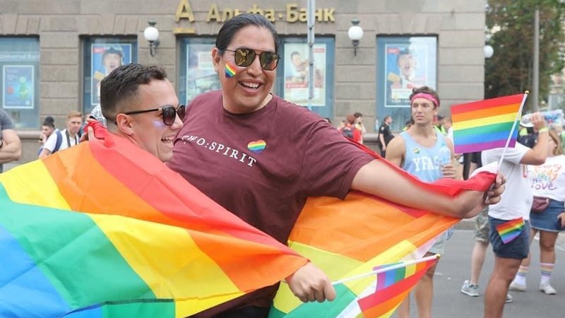 A man with a Native background wraps a rainbow flag around himself and a friend, smiling.