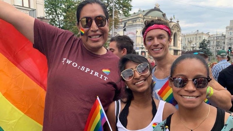 A group of friends huddle together for a picture, smiling, while waving rainbow Pride flags.