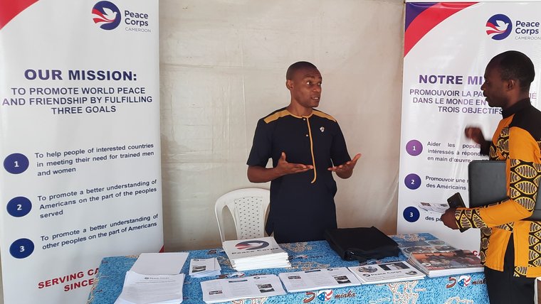 A male person stands in front of a table, talking to another male person.